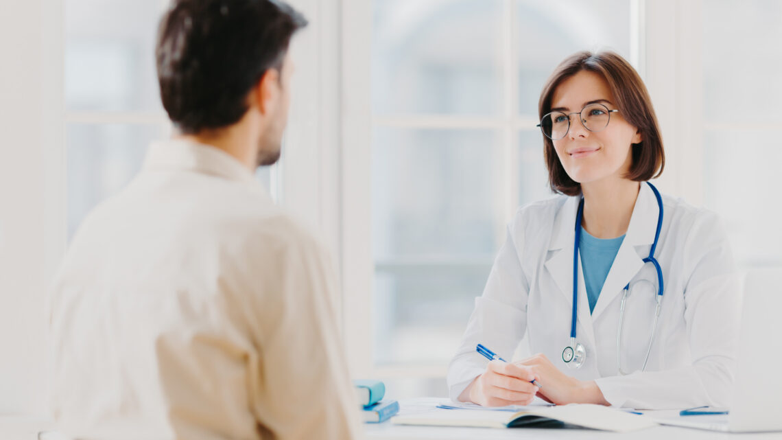 Doctor and patient discuss something, sit at table in clinic Female cardiologist in eyewear gives medical consultation diagnostic, advice for man how to cure disease, pose in hospital room