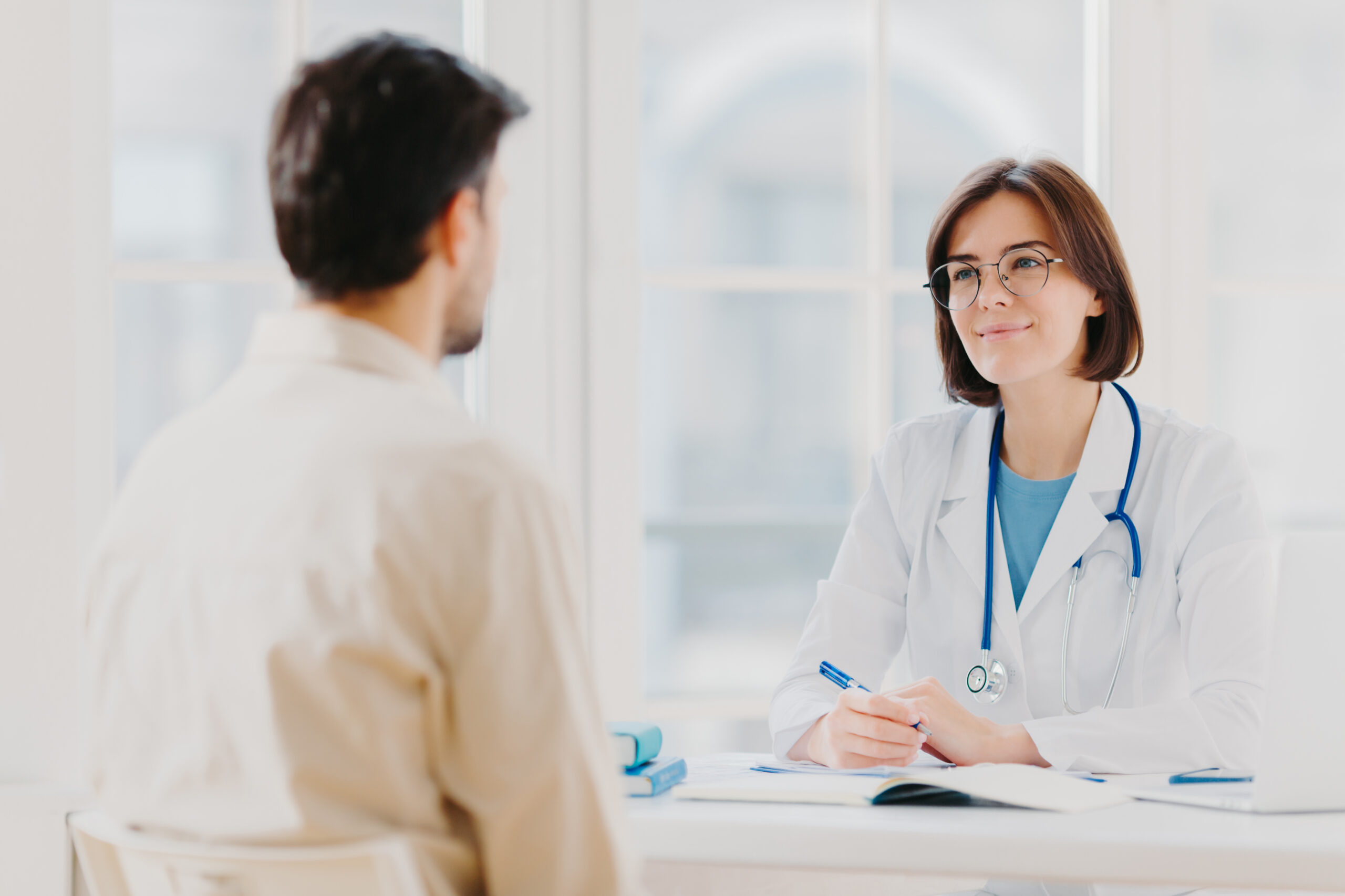 Doctor and patient discuss something, sit at table in clinic Female cardiologist in eyewear gives medical consultation diagnostic, advice for man how to cure disease, pose in hospital room