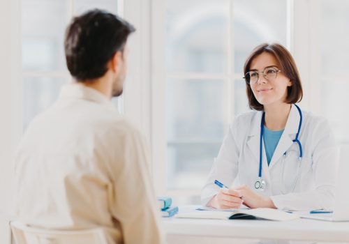 Doctor and patient discuss something, sit at table in clinic Female cardiologist in eyewear gives medical consultation diagnostic, advice for man how to cure disease, pose in hospital room